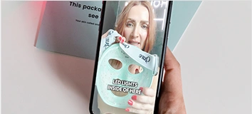 Woman holding phone with LED face mask on screen.